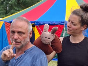 Zwei Menschen und eine Handpuppe vor einem Zirkuszelt