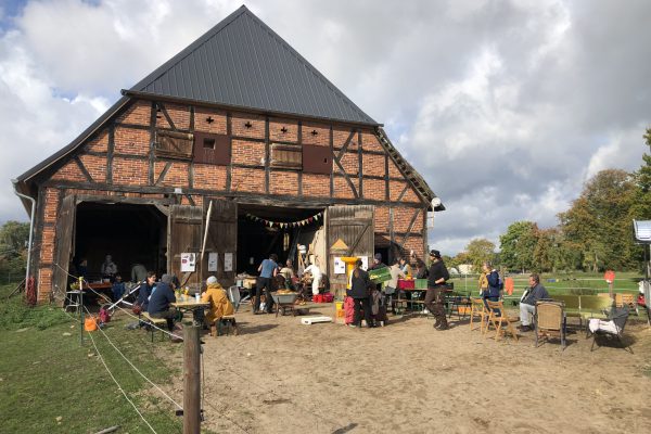 Menschen sitzen unter einem Tarp vor einer alten Scheune